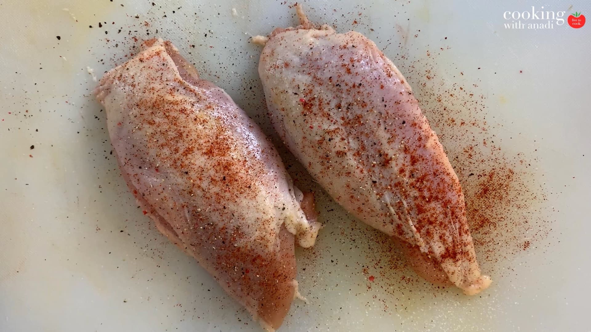 Two seasoned chicken breasts on a light surface, generously coated with salt, pepper, and cayenne pepper.