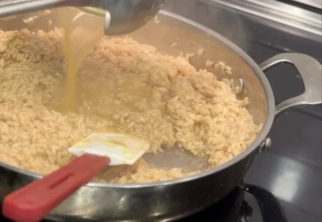 Creamy risotto being stirred in a pan with a red spatula, showing the final stages of cooking with a rich, pale yellow sauce coating the rice grains.