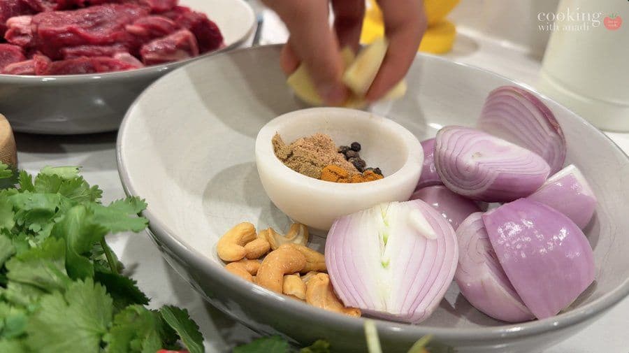 Spice paste ingredients in white bowl with cashews, sliced red onions, and fresh cilantro arranged on plate.