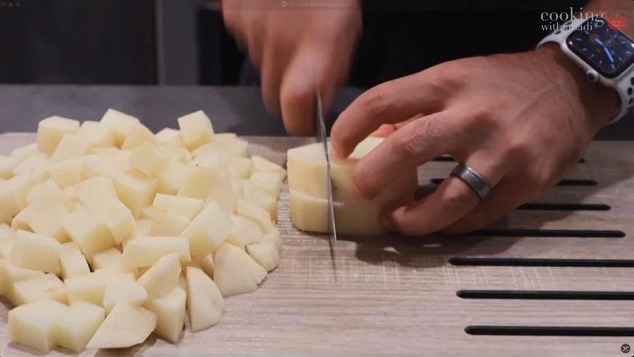 Cubed potatoes on a cutting board with a hand holding a knife, mid-cutting process.