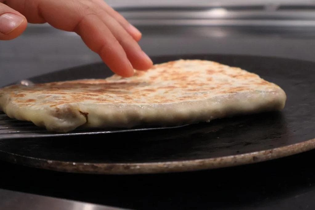 Golden-brown beef martabak cooking in a cast iron pan, being flipped with fingers visible above.