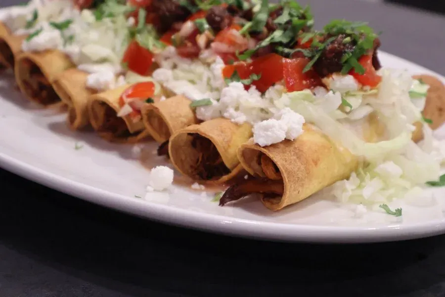 Crispy golden taquitos on a white plate topped with shredded lettuce, diced tomatoes, cilantro, and crumbled cotija cheese.