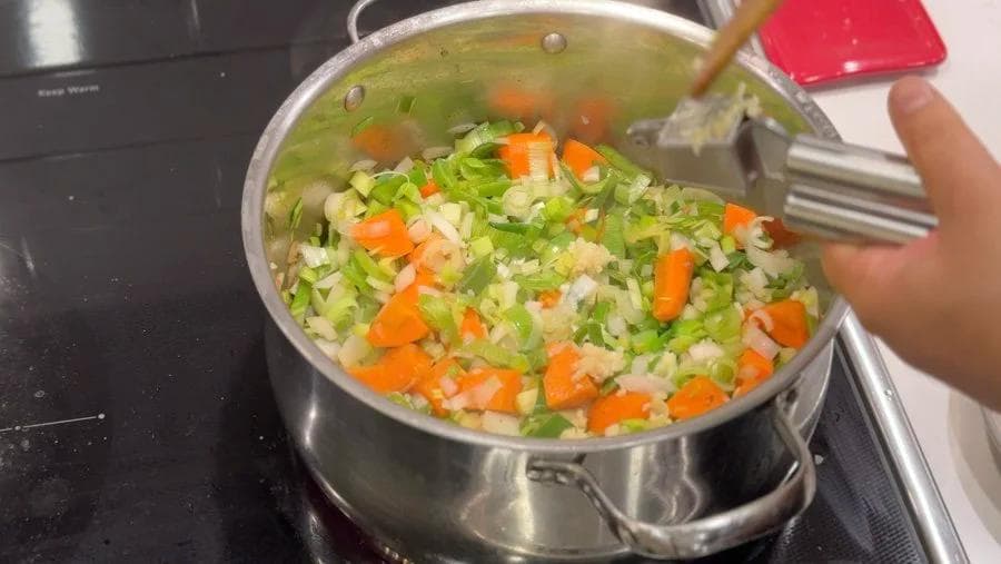 Sautéed onions, carrots, leeks, and garlic in a pot with broth, being stirred with a wooden spoon.