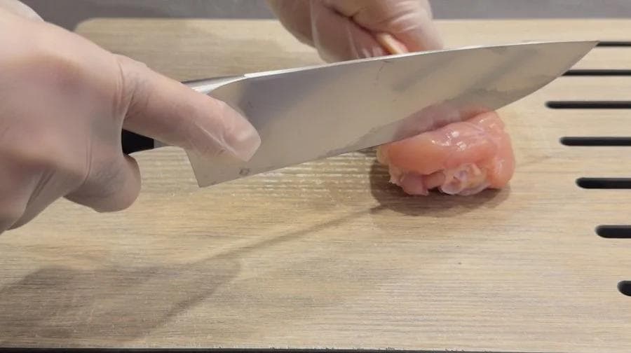 Chef making deep cuts into raw chicken drumstick with sharp knife on wooden cutting board