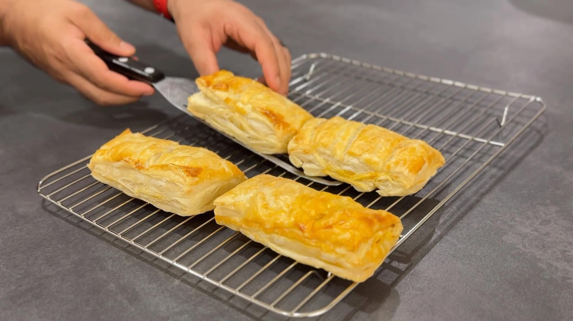 Golden-brown chicken keema puff pastries cooling on a wire rack with hands arranging them.