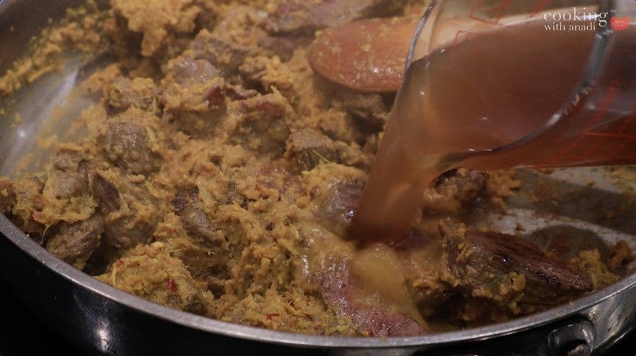 Coconut milk being poured into a pan of cooked beef rendang paste with spices and meat.