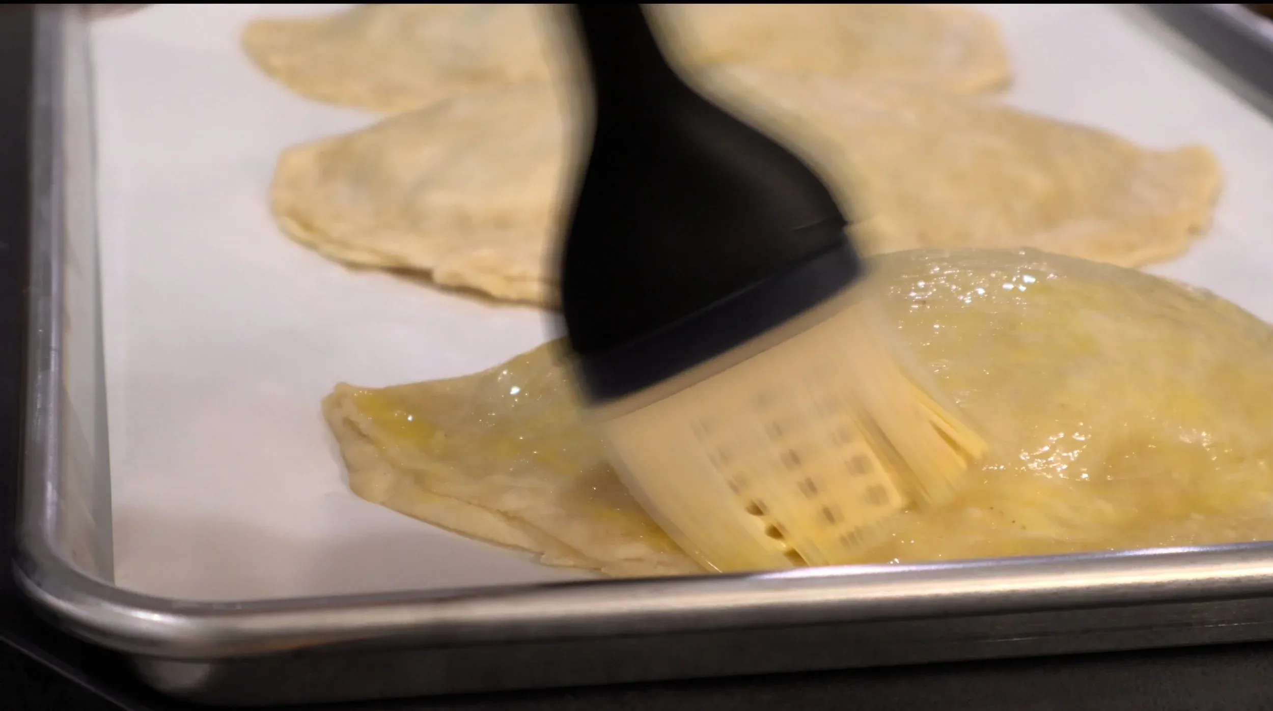 Brushing egg wash onto unbaked hand pies with a pastry brush on parchment paper.