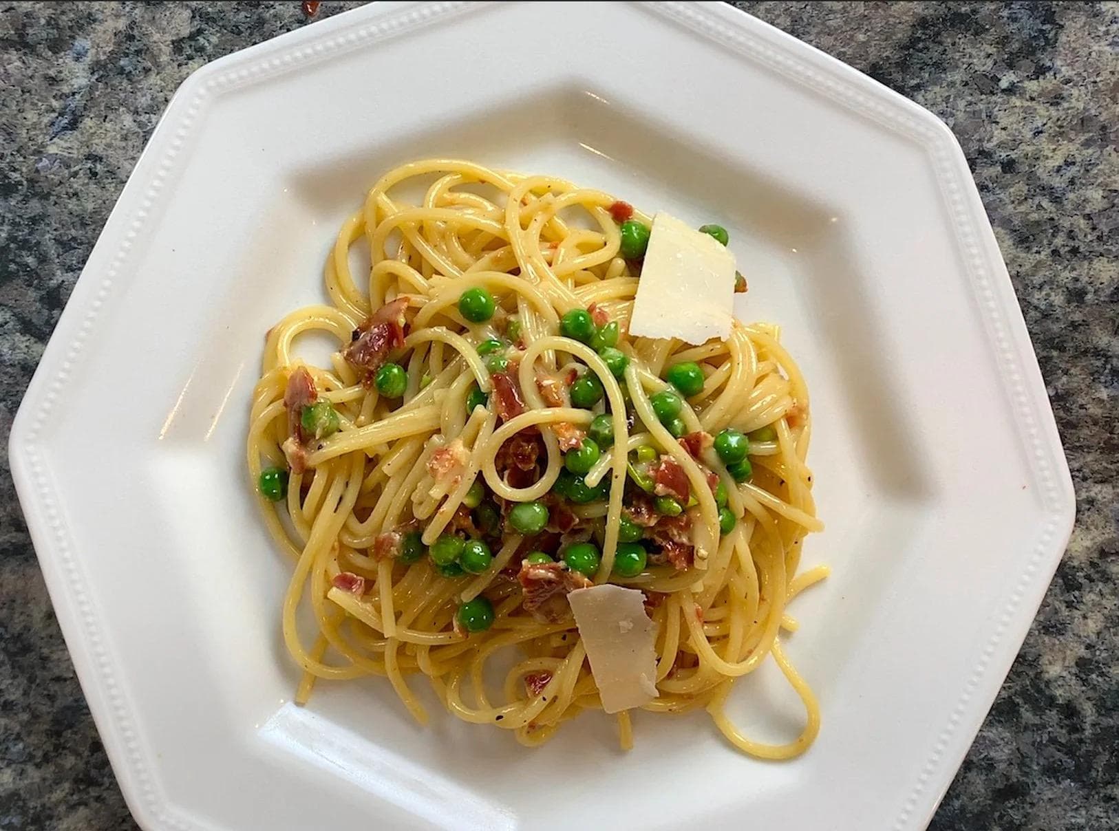 Spaghetti alla carbonara on a white plate with creamy sauce, peas, bacon, and shaved parmesan cheese.