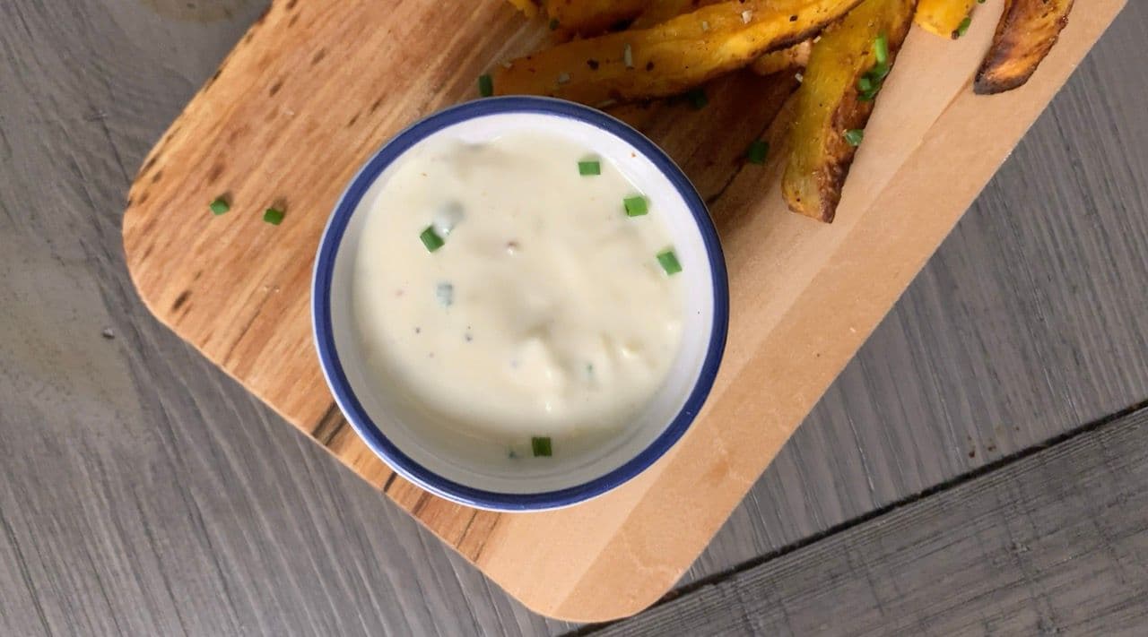 Garlic truffle aioli in a blue-rimmed bowl with chive garnish, served with crispy potato wedges on a wooden board.