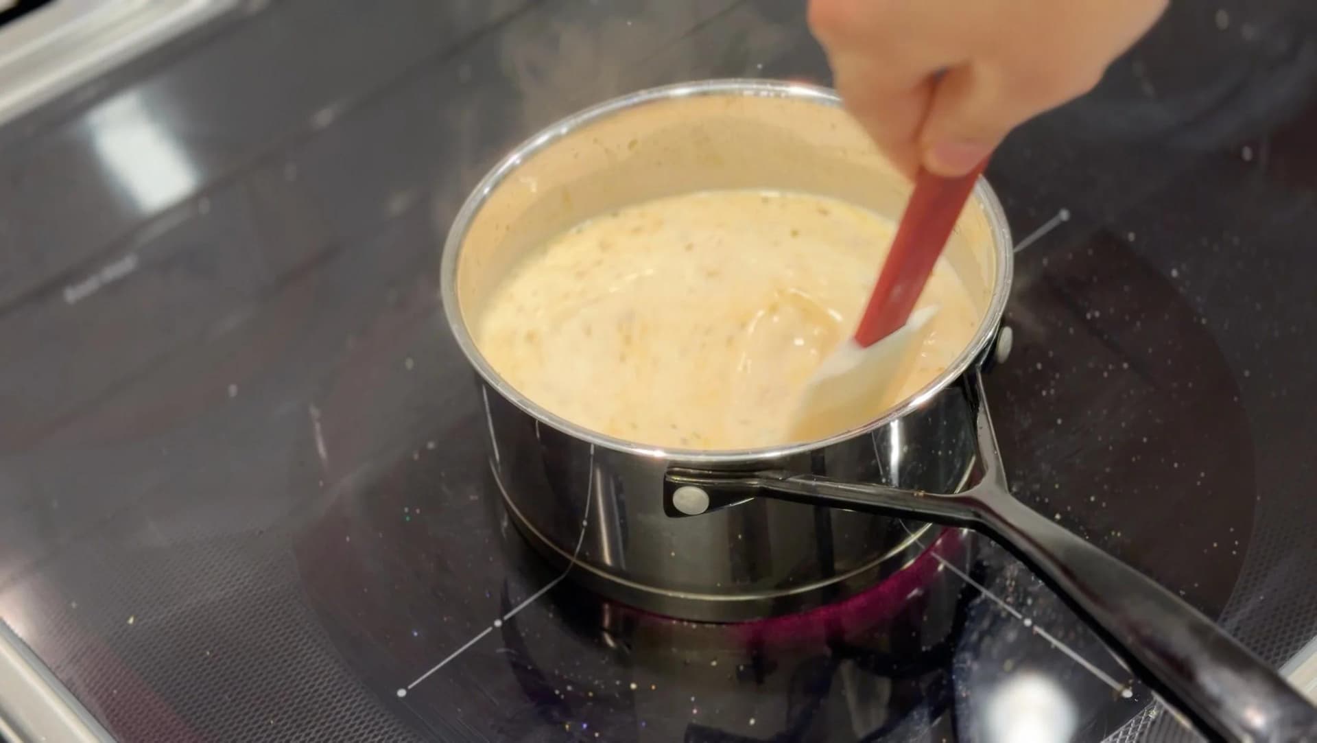 Whisking creamy béchamel sauce in a saucepan on the stovetop with a red whisk.