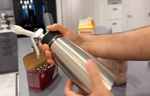 Hand holding a stainless steel whipped cream dispenser with black head and trigger over a kitchen counter.