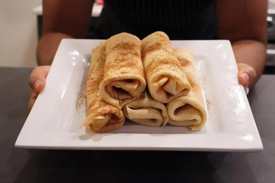 Five rolled pumpkin crepes dusted with cinnamon on a white square plate held by hands.