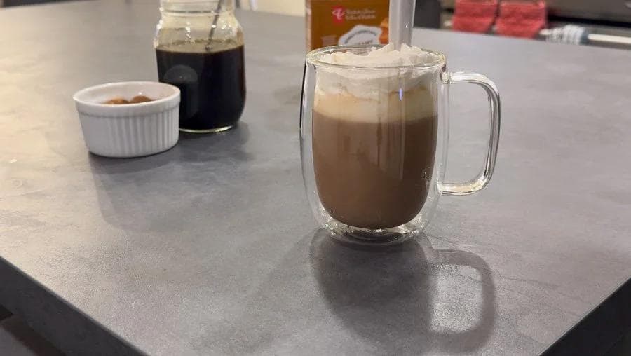 Gingerbread latte in a clear glass mug topped with whipped cream, displayed on a gray countertop with coffee ingredients nearby.