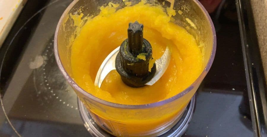 Food processor blending cooked pumpkin into smooth orange puree with water added.