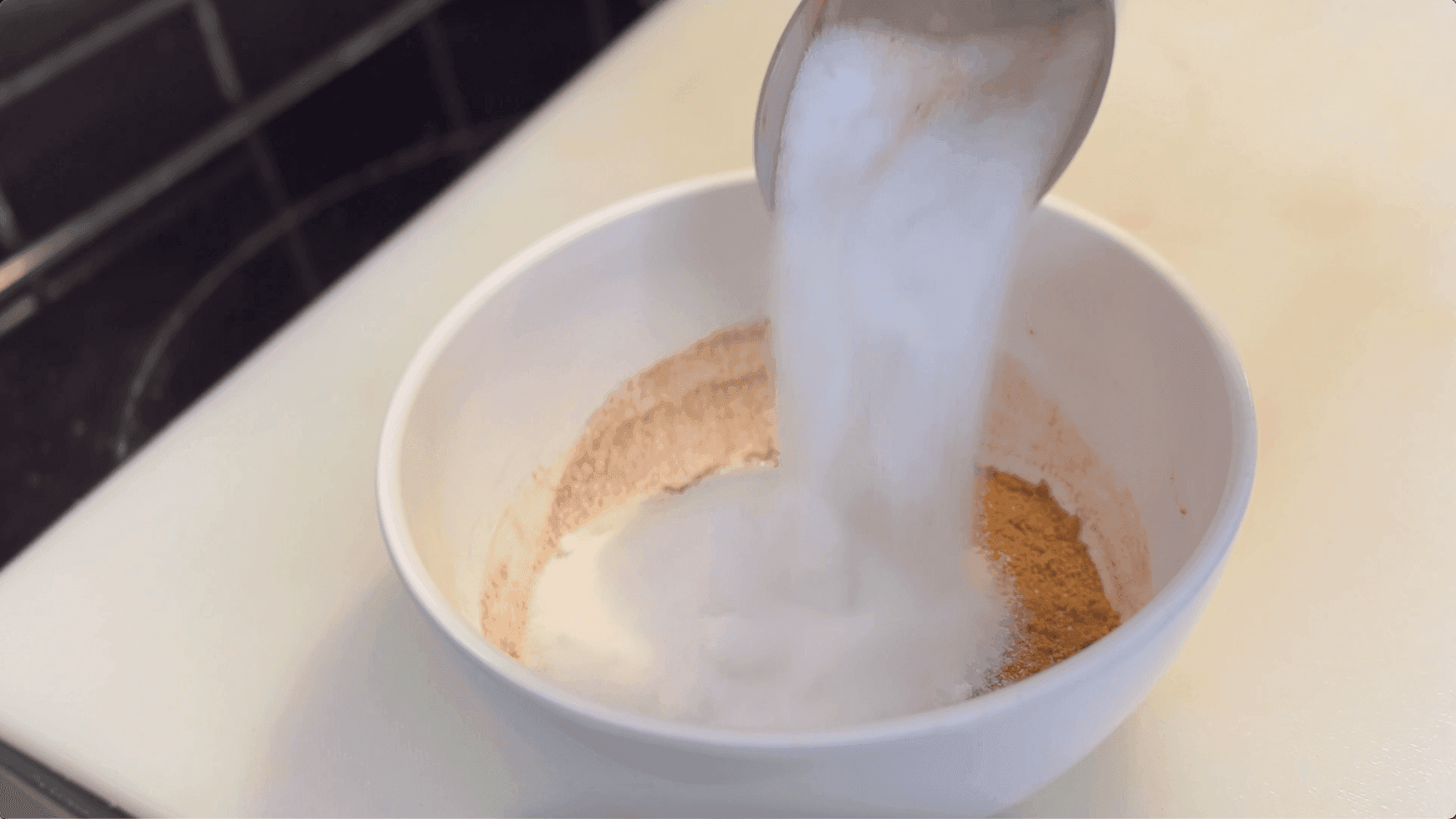 Pouring sugar mixture into a white bowl containing cinnamon and brown sugar for cinnamon roll filling.