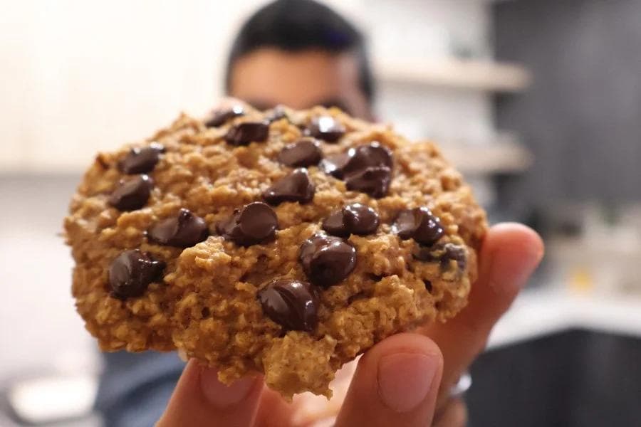 Hand holding a thick pumpkin chocolate chip cookie with chocolate chips throughout, blurred kitchen background