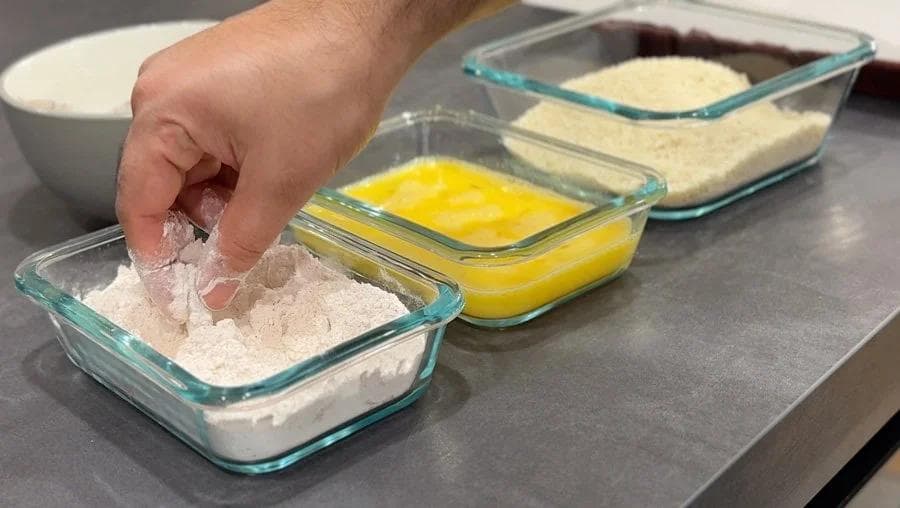 Hand dipping shrimp in flour coating station with eggs and breadcrumbs in separate glass bowls.