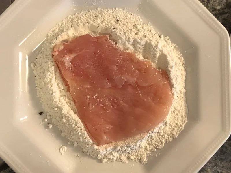 Raw chicken breast dredged in flour with egg coating, ready for panko breading in white bowl.