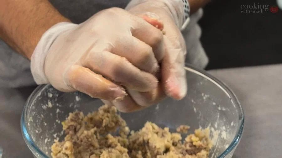 Hands in plastic gloves mixing ground potato and meat mixture in a glass bowl.