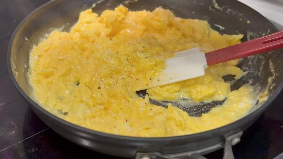 Scrambled eggs cooking in a pan with a red spatula, seasoned with black pepper