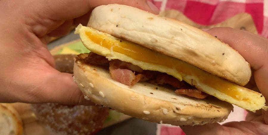 Hands holding an assembled bagel sandwich with egg, bacon, and melted cheese between toasted bagel halves.