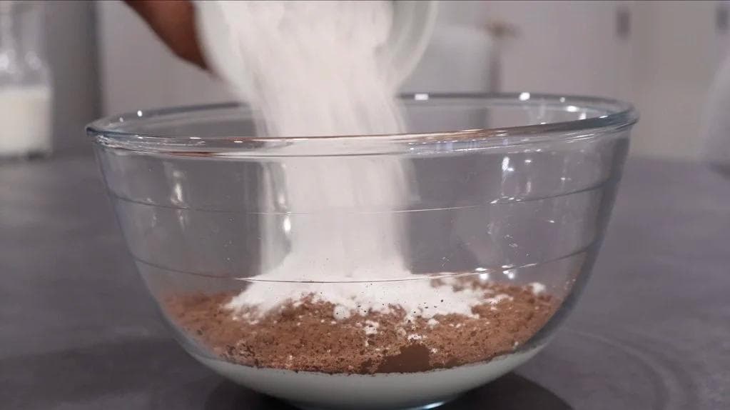 Cocoa powder and sugar being poured into a glass bowl containing heavy cream for whipped cream.
