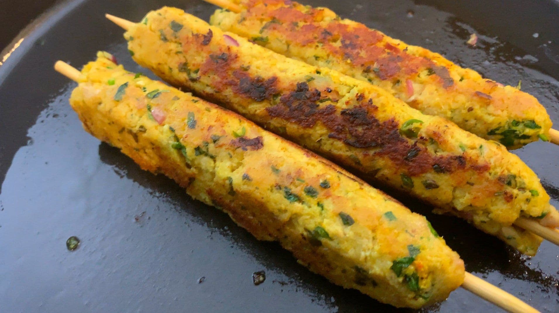 Three juicy Indian chicken seekh kebabs on skewers cooking on a dark griddle with charred spots and fresh herbs.