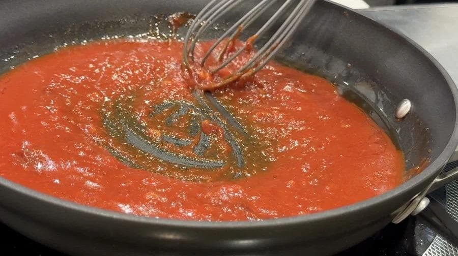 Red sauce simmering in a dark pan with a whisk mixing in seasonings.