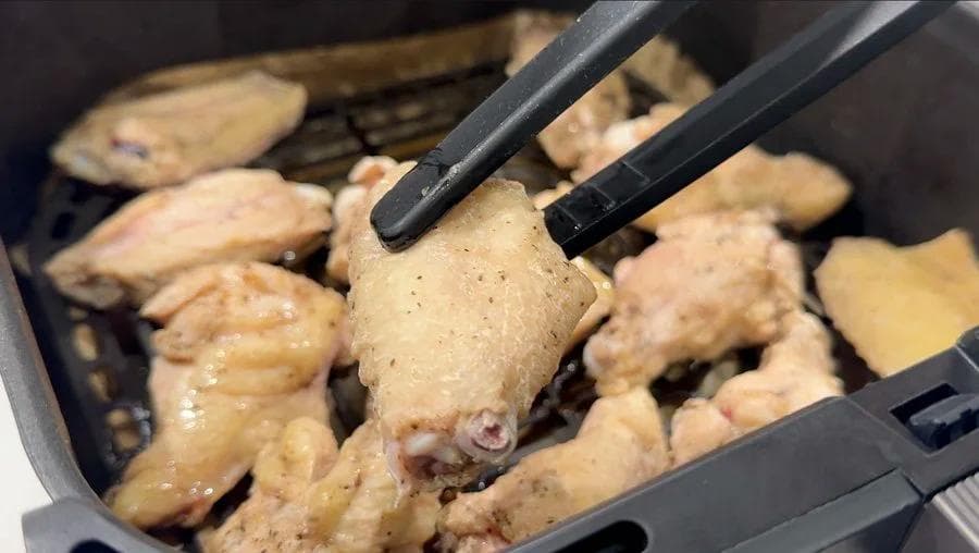 Tongs lifting a partially cooked chicken wing from an air fryer basket filled with wings in various stages of cooking.
