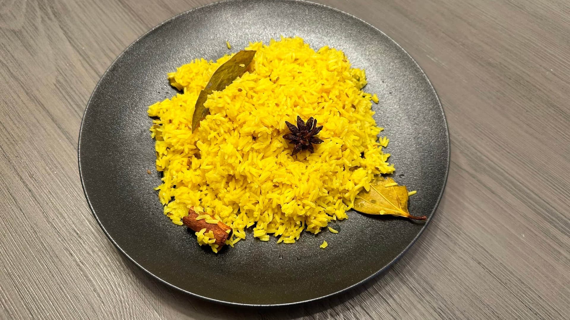 Fluffy turmeric rice served on a dark plate, garnished with bay leaf, star anise, and cardamom pods.