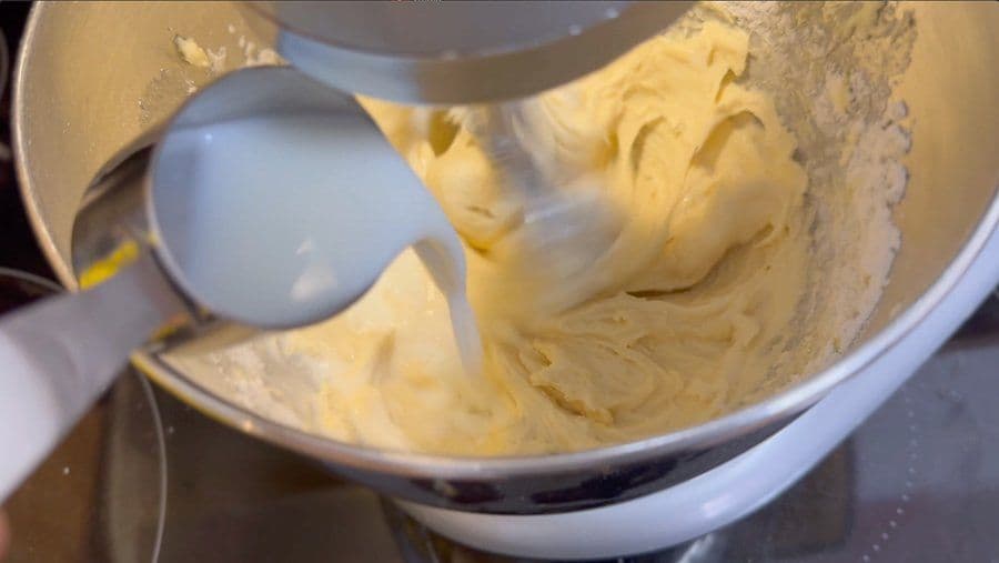 Pouring milk into thick yellow cookie batter in a mixing bowl with a hand mixer.