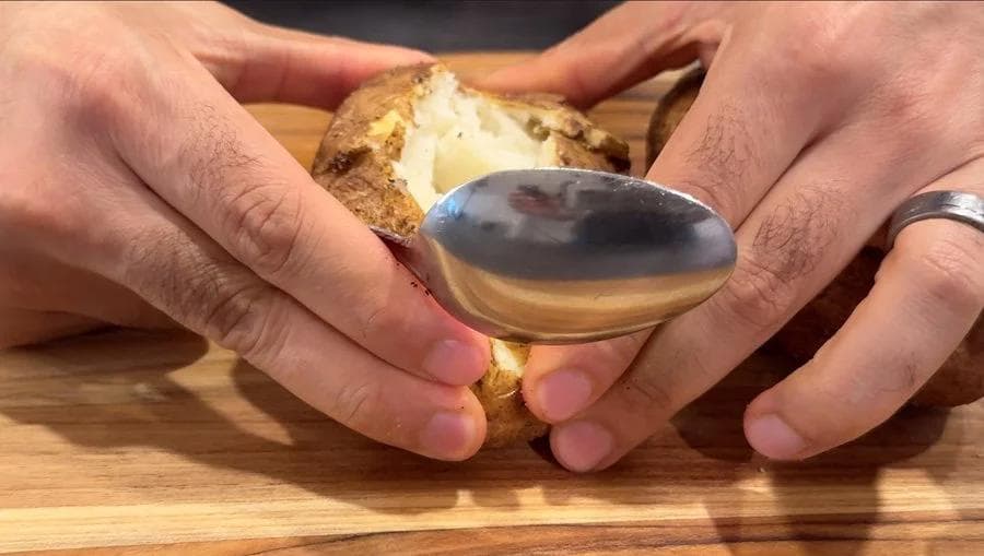 Hands scooping fluffy potato interior from a halved baked potato skin using a spoon on a wooden surface.