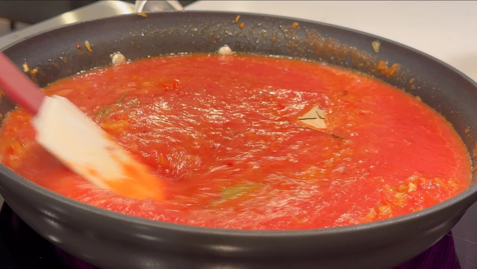 Tomato sauce simmering in a dark skillet, being stirred with a red-handled spatula during piperade preparation.
