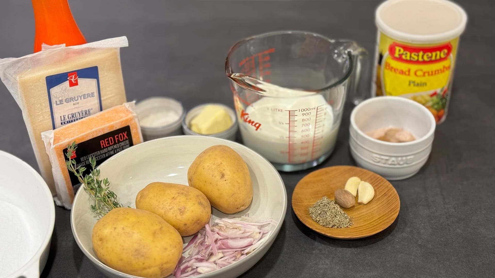 Ingredients for potato gratin arranged on a dark surface: Yukon gold potatoes, shallots, butter, garlic, cream, cheese, and fresh thyme.