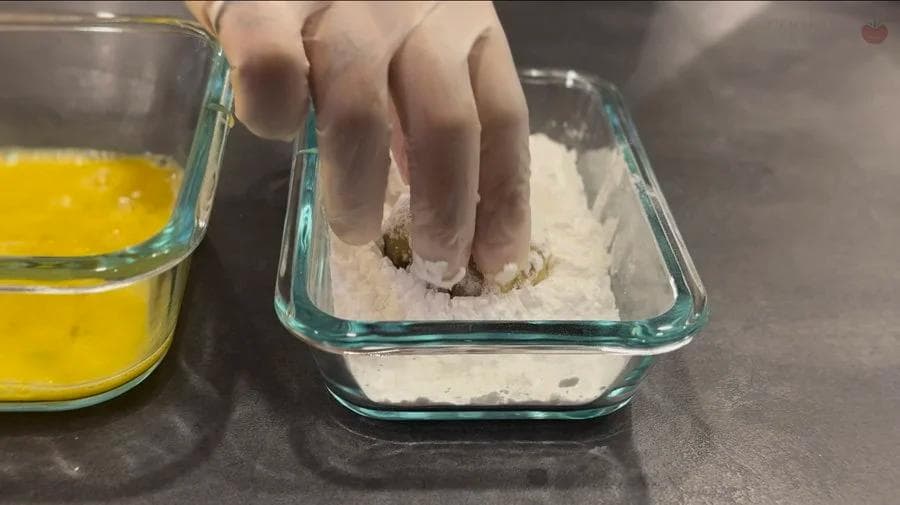 Hand coating potato fritter patty in flour mixture in glass baking dish next to bowl of beaten egg.
