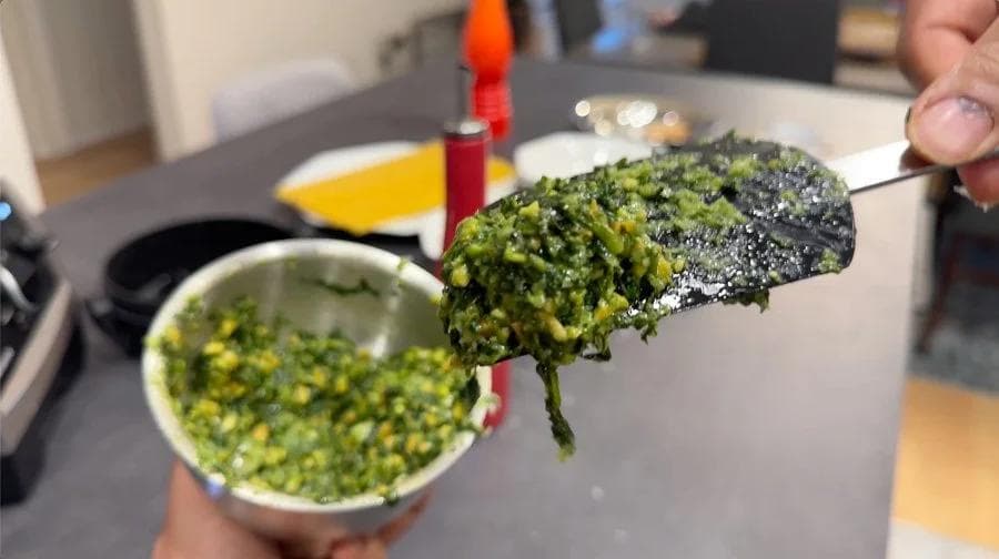 Creamy green pesto mixture on a spoon held over a mortar bowl, showing coarse paste consistency.