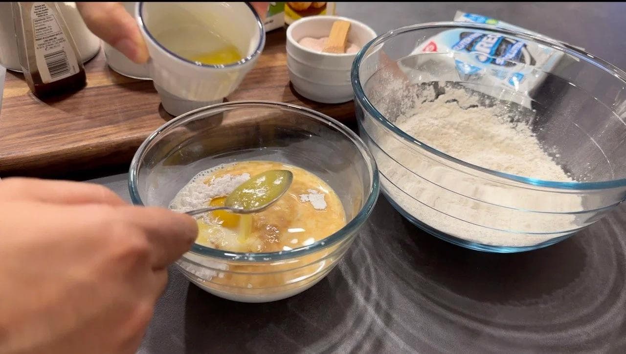 Wet ingredients being stirred in a glass bowl with a spatula next to a bowl of dry flour mixture on a dark surface.