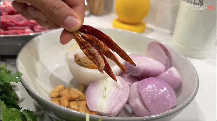 Hand holding dried red chilies over a white bowl containing sliced red onions, garlic, and cashews for rendang spice paste.