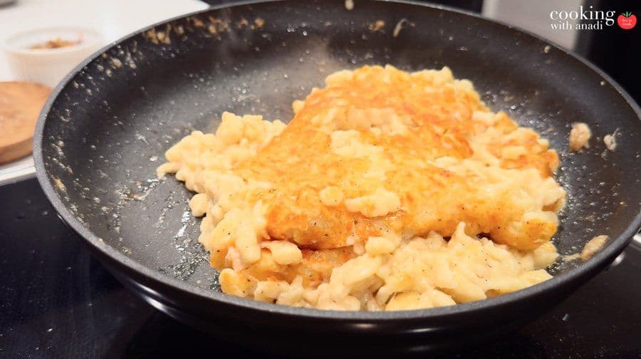 Käsespätzle with melted cheese folded in, showing a golden-brown crust forming on the bottom in a dark skillet.
