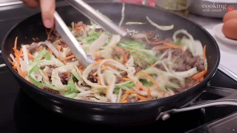 Stir-frying cabbage, carrots, and meat in a black skillet with green onions and sauce being mixed together.