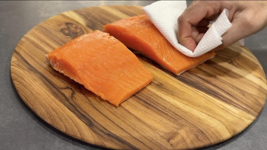 Salmon fillets being patted dry with paper towels on a wooden cutting board.