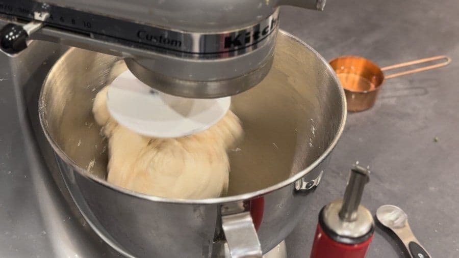Stand mixer with dough hook kneading pizza dough in metal bowl, soft and slightly sticky dough visible.
