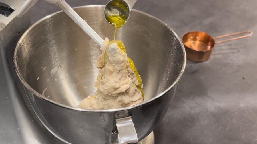 Olive oil being drizzled from a spoon onto pizza dough in a stainless steel mixing bowl.