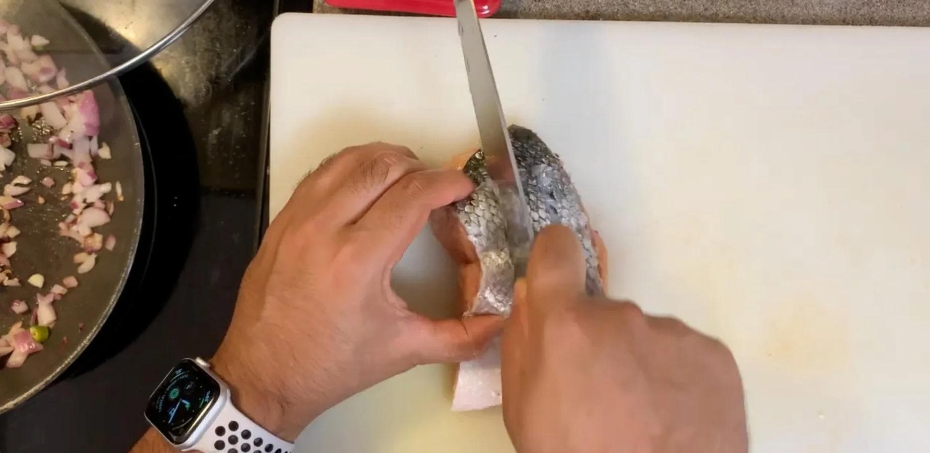 Hands descaling salmon fillet with a knife on a cutting board, with a pan of ingredients visible in background.