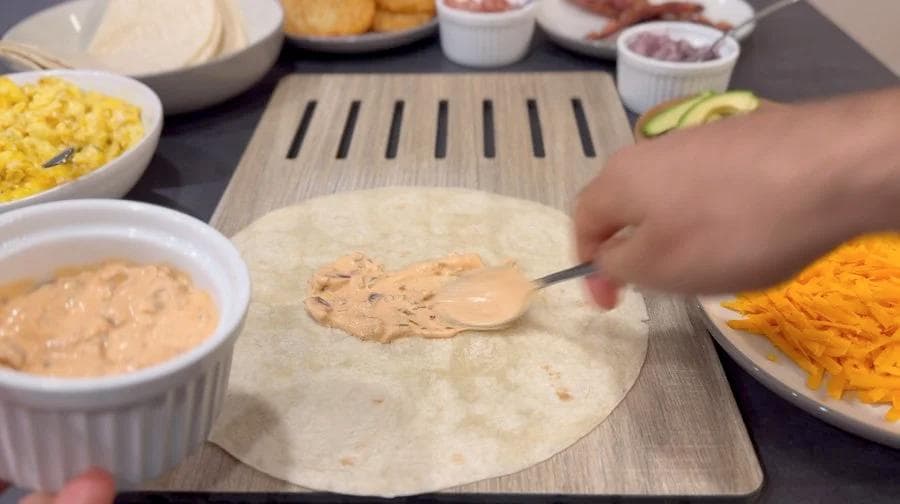 Spreading burrito sauce on a flour tortilla with ingredients like scrambled eggs, cheese, and hash browns nearby.
