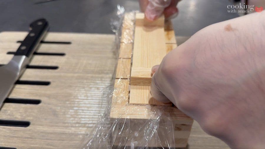 Pressing down on oshizushi mold with plastic wrap covering the sushi to compact it firmly.
