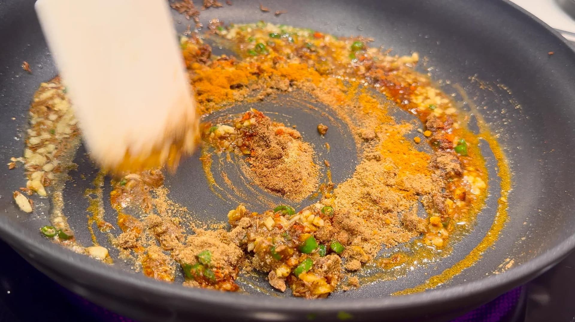 Spiced potato and pea mixture being stirred in a dark pan with a spatula, showing cooked spices and vegetable pieces.