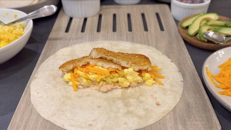 Breakfast burrito assembled on a tortilla with scrambled eggs, cheddar cheese, and hash browns between toasted bread slices.