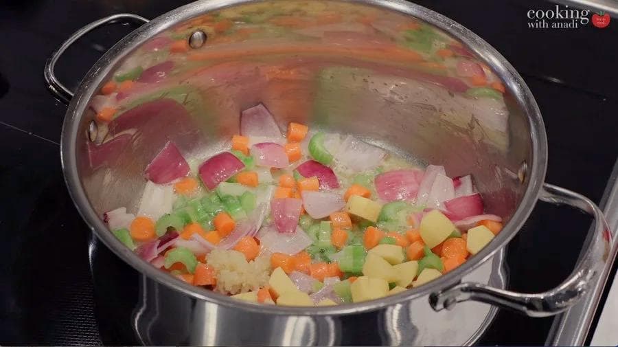 Diced vegetables including onions, carrots, celery, potatoes, and peas cooking in a large pot over heat.