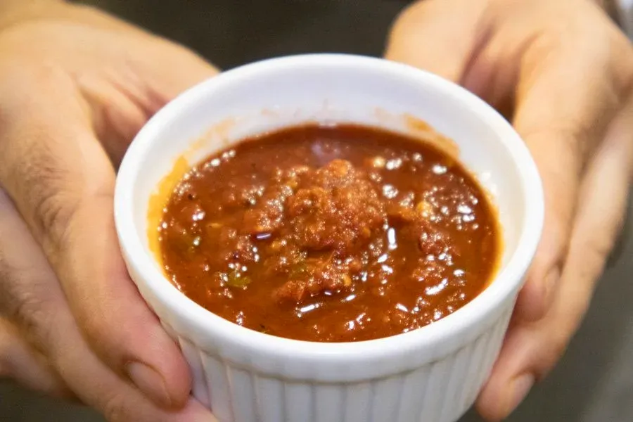 Hands holding a white ramekin of homemade salsa roja with a glossy, chunky texture